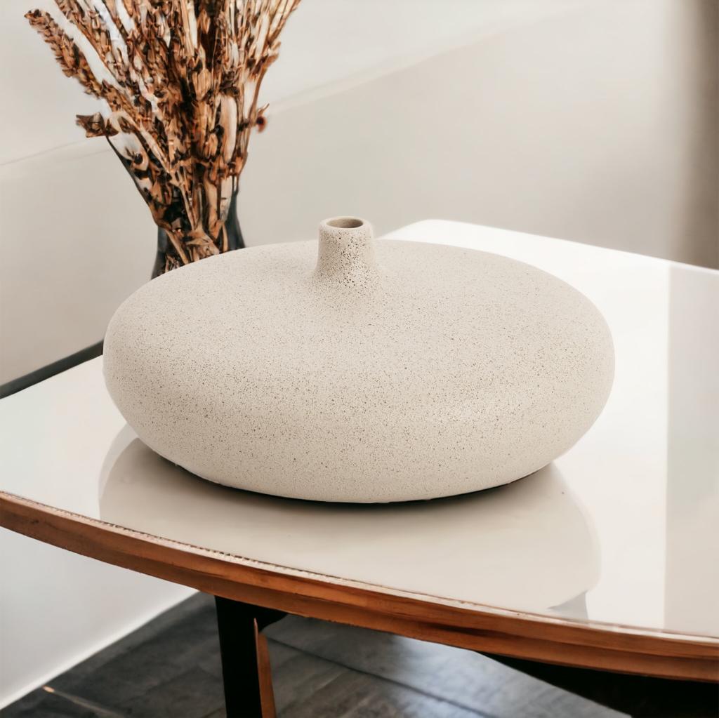 Vaso de cerâmica com design moderno achatado e uma abertura central pequena, revestido em uma textura granulada neutra. Sua forma baixa e ampla é inspirada na estética minimalista, tornando-o perfeito para decorações contemporâneas ou de inspiração escandinava. Este vaso é uma escolha excelente para quem procura peças decorativas sutis, mas impactantes, para complementar qualquer espaço interno com um toque de sofisticação e design limpo.