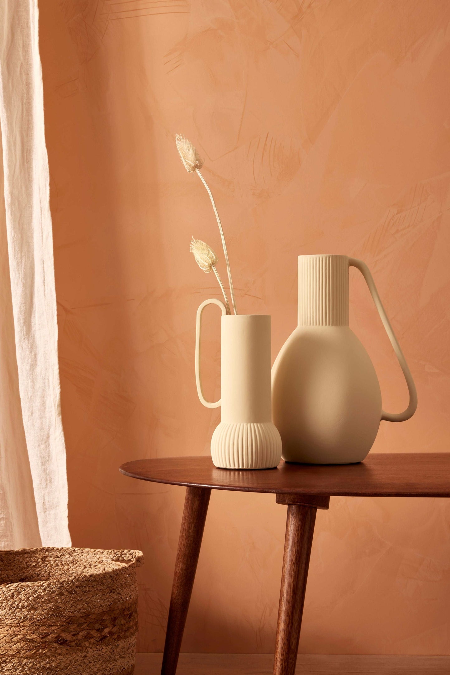 Cena de interiores com um fundo de parede cor de pêssego texturizado. Sobre uma mesa redonda de madeira escura, estão dois vasos cerâmicos: um alto e esbelto na cor creme com detalhes canelados na base e uma alça, contendo duas flores secas de tonalidade clara; o segundo, mais volumoso com um design arredondado e uma alça proeminente. À esquerda, um pedaço de tecido branco pendurado e, na parte inferior esquerda, é visível a borda superior de uma cesta tecida. O vaso menor é o Gruna e o grande o lotus